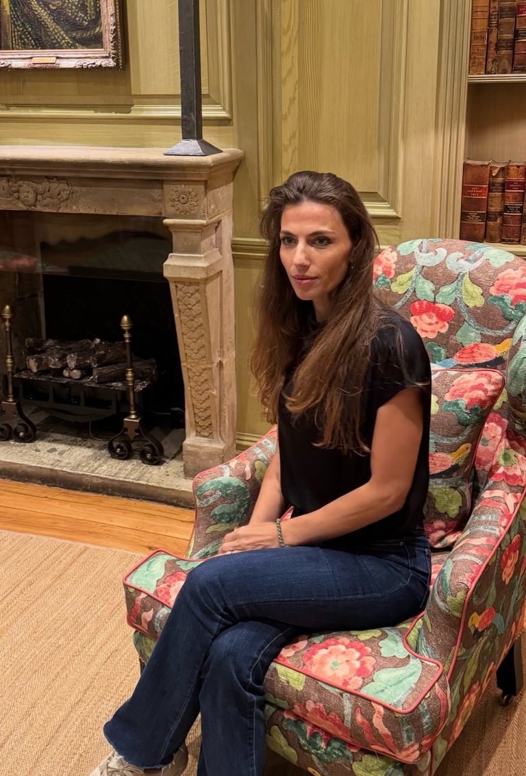 Woman with long brown hair sitting in a floral patterned armchair in an elegant room with a fireplace and bookcase